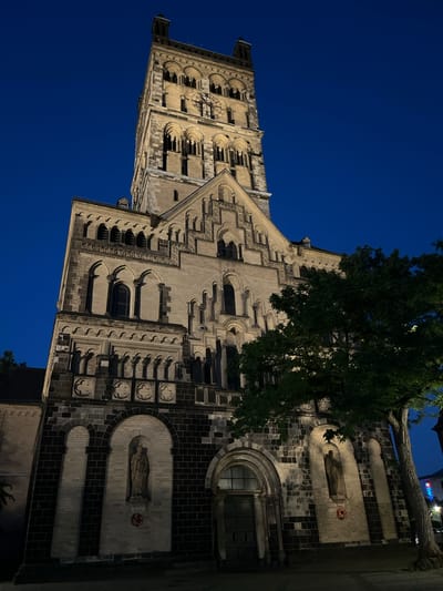 The image depicts the St. Quirinus Minster in Neuss, Germany, viewed from a low angle at dusk. The Romanesque basilica, constructed from light-colored stone and dark brick, features a tall, imposing tower with arched windows and a clock face. The façade is adorned with intricate architectural details, including rows of small arches and decorative carvings. Two niches on the lower level contain statues of religious figures. A large, leafy green tree partially obscures the right side of the building. The sky is a deep, clear blue, indicating it is nighttime. The scene is illuminated by artificial lighting, casting shadows and highlighting the textures of the stone. There are no people visible in the frame, and the environment appears to be an urban square or street.