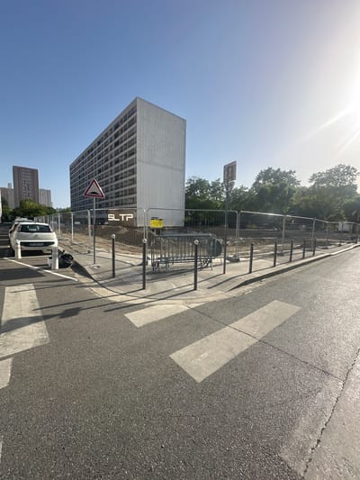 The image depicts a street scene in Villeurbanne, France, on a sunny day. In the foreground, a crosswalk is visible on an asphalt road, with shadows cast by nearby objects. To the left, a white car is parked near a sidewalk. A tall, modern building with numerous windows dominates the background, and a smaller, older building with red brickwork is visible further to the left. The area behind the sidewalk appears to be a construction site, enclosed by temporary metal fencing. A sign with the letters "SLTP" is prominently displayed on the fence, indicating a construction company. Several metal barriers and bollards are also present along the sidewalk. Trees with green foliage are seen behind the construction site, and the sky is clear and blue.