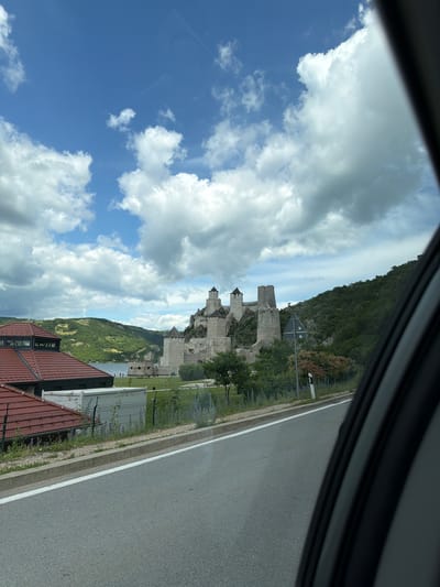 The image, taken from a vehicle, shows a view of the Golubac Fortress in Golubac Municipality, Serbia. The fortress, constructed of stone, is situated on a rocky outcrop overlooking a body of water. It features multiple towers and walls, some with dark-colored roofs. In the foreground, a paved road with a white line runs alongside a grassy area and a fence. To the left of the road, a building with a red, tiled roof is visible. The background consists of a green, tree-covered hillside and a cloudy blue sky. Several wind turbines can be seen on the distant hills.