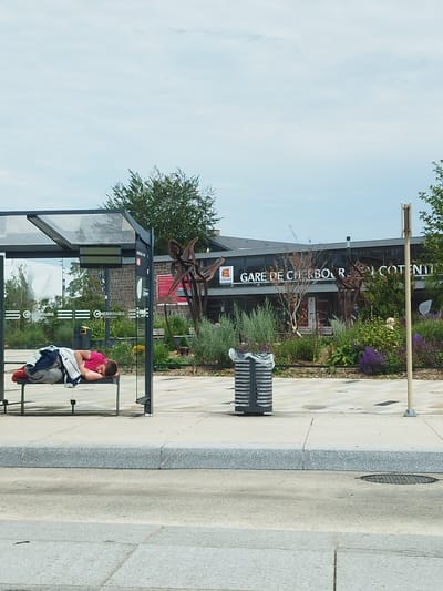 A person is sleeping on a bench at a bus stop in Cherbourg-en-Cotentin, France. The bus stop has a glass shelter and metal supports. Behind the bus stop is a building with the text "GARE DE CHERBOURG en COTENTIN" visible. The person is wearing a pink shirt and grey pants, and is covered with clothing. There are plants and flowers in front of the building. A trash can is located to the right of the person. The sky is cloudy.