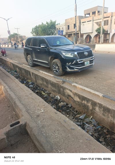A black SUV, likely a Toyota Land Cruiser Prado, straddles a concrete drainage ditch filled with litter. The vehicle faces right, partially blocking the road. Buildings line the background, some with arched openings. Other vehicles and people appear further down the road. The scene appears to be an urban area with visible signs of pollution. The date in the corner says 2026.01.28 07:47 AM.