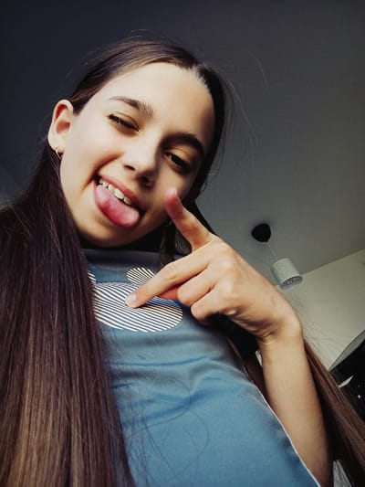 A young female with long brown hair is shown from a low angle, looking up at the camera. She is winking and sticking her tongue out, with her right index finger pointing towards the camera. She is wearing a blue t-shirt with a white Mickey Mouse-like graphic on it. The background consists of a ceiling with a pendant light fixture. The image appears to be a selfie taken indoors. The location is identified as Radzymin, Poland.