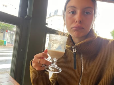 A young adult female is depicted in a close-up, selfie-style indoor portrait, located in Deauville, France. She has light skin, dark hair, and a neutral expression with slightly pursed lips, looking directly at the camera. She is wearing a mustard yellow, ribbed, high-collared sweater with a prominent silver-toned zipper and a black leather pull tab. Her right hand, positioned in the lower-center, holds a clear glass goblet containing remnants of a light brown, frothy beverage. A thin silver-toned ring is on her right ring finger, and a darker, patterned ring is on her right middle finger. The interior foreground includes the edge of a light brown wooden table. A dark green window frame divides the interior from the exterior view. Outside, a street features a white-striped crosswalk. A green public trash can and a red traffic light are visible on the sidewalk. Across the street, various storefronts and buildings with typical European architecture are present under an overcast sky.
