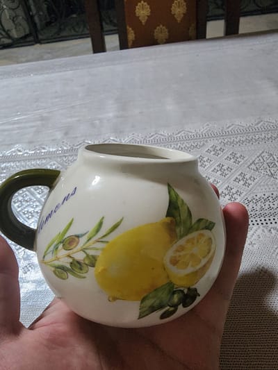 The image shows a white ceramic pitcher with a green handle decorated with lemons and olive branches. The word "Lemons" is written in blue script on the pitcher. A person's hand is holding the pitcher, which is placed on a white tablecloth with a lace-like pattern. The background is out of focus, but appears to be an indoor setting with furniture and possibly a window. The city of Gudermes, Russia, is not mentioned or depicted in the image.