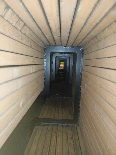 An interior view looking down a tunnel-like structure made of wooden planks and framed by black metal. The structure appears to be a passage or walkway with a series of nested rectangular openings leading to an exit in the distance where a glimpse of green and sky is visible. The wooden planks are oriented lengthwise along the tunnel, creating a sense of depth and perspective. The lighting is dim inside the tunnel, with the brightest light emanating from the exit. There are no people or specific actions depicted in the image. The scene suggests an architectural element possibly found in a park, outdoor recreational area, or a unique passage in an urban setting. The provided location, Radkevshchina, Russia, cannot be verified from the image alone.