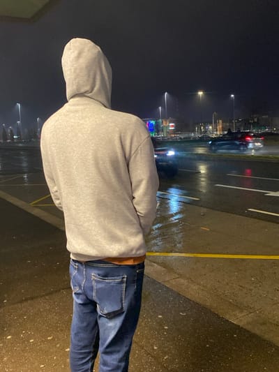A person stands on a wet platform at night. The individual wears a gray hooded sweatshirt and dark jeans. Light reflects off the wet pavement, showing street markings and a yellow safety line. Streetlights and buildings are visible in the background, with blurry light trails from cars. The scene suggests a rainy, urban environment.