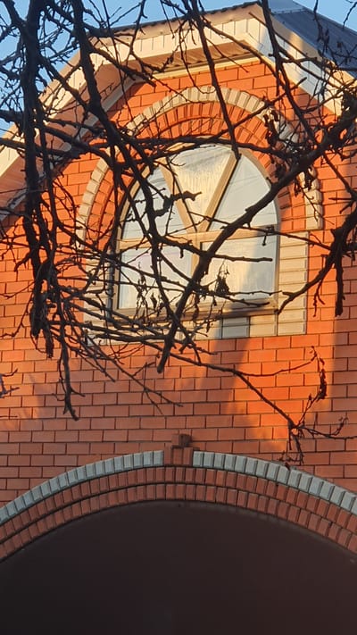The image shows the upper facade of a brick building, possibly a house, with an arched window. The brickwork is predominantly orange-red, with a decorative arch above the window featuring lighter colored bricks. The window itself is framed by a wooden structure and appears to have a frosted or textured glass. Bare tree branches with no leaves obscure parts of the facade and window. The lighting suggests it is either sunrise or sunset, casting a warm glow on the bricks. The sky above the building is a clear blue. The image is taken from a low angle, looking upwards. The location is identified as Ust-Labinsk, Russia.