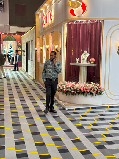 An adult male is captured mid-stride in an indoor commercial or exhibition space, likely within Jaipur Municipal Corporation, India. He appears approximately 30-40 years old, wearing a green and white patterned long-sleeved shirt, dark trousers, a black belt with a silver buckle, and dark shoes. A lanyard is visible around his neck. He holds a black smartphone to his right ear, speaking on a call, with his left hand in his left pocket. He looks towards the right side of the frame, exhibiting a slight smile.
The floor is covered by a patterned carpet featuring light gray, dark gray, and yellow rectangular blocks arranged in a horizontal grid. To the man's right, a prominent storefront is visible, labeled "Saanre BY SAANRE CREATIONS PVT LTD," suggesting a jewelry or luxury goods establishment. The store's exterior is characterized by white and gold paneling, with framed sections displaying artwork or promotional material and multiple wall sconces emitting warm light. Above the store's name, a distinctive red and gold abstract logo is affixed.
A large display window features deep red velvet curtains as a backdrop. Inside, a white pedestal holds a prominent green and white gemstone necklace displayed on a bust, flanked by a pair of matching earrings. A small pink floral arrangement is also present on the display. Below the display window, a white, curvilinear planter is filled with numerous pink roses and white filler flowers. In the background, other sections of the exhibition hall or additional storefronts are visible, including one with arched doorways and draped fabric. Several other individuals are seen further down the corridor. The area is well-lit by artificial lighting.