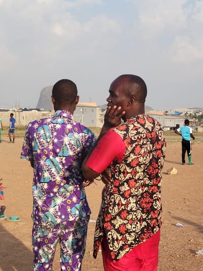 Two men are standing in a dirt clearing outside an urban area. One man is wearing a red-and-black patterned shirt with red sleeves and is touching his face. The other man is wearing a purple shirt with a light blue-and-white floral pattern. In the background are other people, buildings, and a hillside. The location is Ibo, Nigeria.