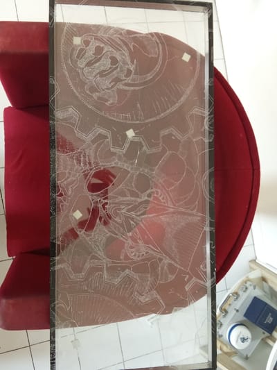 A vertically oriented rectangular glass panel, approximately 1:2 aspect ratio, with a black frame, is centrally positioned. The panel is etched or frosted with a detailed, intricate design featuring interconnected gears, cogwheels, and stylized organic-mechanical forms, including a brain-like element in the upper portion. Four small, white square adhesive markers are affixed to the etched surface. The glass exhibits multiple fractures and cracks, concentrated along its lower and left edges, with some apparent minor damage to the black framing.

The panel is situated over a deep red upholstered armchair or sofa section, which provides a contrasting background. A reflection of a human hand is discernible on the glass surface, predominantly in the lower-central area. The lower right portion of the scene reveals a light-colored tiled floor with dark grout lines. Resting on a low, light-colored wooden stand in this area are a silver-colored case with a handle, a dark blue rectangular box partially labeled 'VALERFELD', and a white jar with a blue lid and a blue logo. The scene is illuminated by ambient light. This location is in Port-Vendres, France.