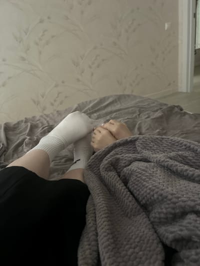 The image depicts a close-up view of two people lying down on a bed, likely in a residential setting in Veshki, Russia. Their legs are extended, and they are covered by a textured grey blanket. One person is wearing white socks and black shorts, while the other is also wearing white socks. Their hands are intertwined, suggesting an intimate interaction. The background shows patterned wallpaper with a light beige tone, and a glimpse of a doorway with white trim is visible to the right, hinting at an adjoining room or hallway. The overall scene is domestic and relaxed.