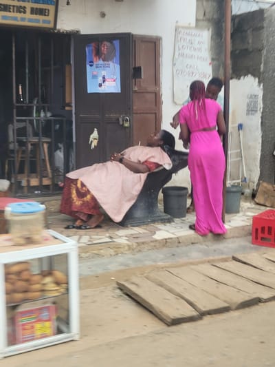 The image depicts a street scene in Katambe, Nigeria. In the foreground, a white display case filled with baked goods sits on the left. A wooden plank pathway is laid across the ground in the middle. Further back, a woman with a pink dress is styling the hair of another woman who is seated in a barber's chair, covered by a striped gown. Behind them, a building has a dark brown door with a poster on it. To the right of the door, a handwritten sign advertises "Readlocks Stylist and Lotions Needed Here. Call 07089229967." Several dark gray trash bins are visible, along with a red plastic crate. The overall environment appears to be an outdoor commercial area.