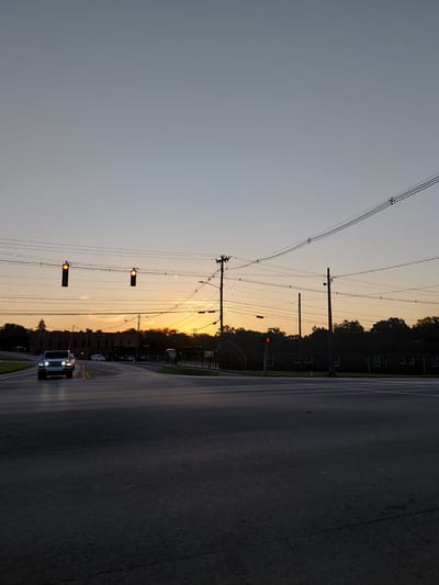 The image depicts a street scene at dusk. The sky is a pale gray, transitioning to a warm orange and yellow hue near the horizon, indicating the sun is setting. Silhouetted against the colorful sky are trees and buildings, with power lines crisscrossing the frame. Two traffic lights, both displaying a red signal, are visible. A white SUV is driving on the asphalt road, its headlights illuminated. The road appears to be a multi-lane street with painted lane dividers. The setting suggests an urban or suburban environment in the United States. The overall mood is peaceful, with the fading light creating a serene atmosphere. No visible text is present in the image.