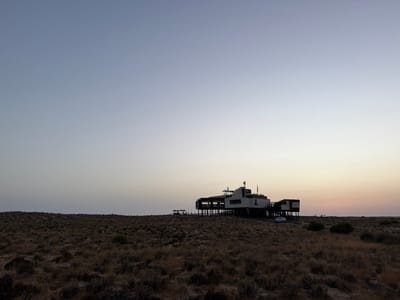 An exterior long shot depicts a modern, multi-level building elevated on numerous dark stilts, situated within an expansive field of low, dry, brownish vegetation. The scene is illuminated by the low sun, indicated by a sky transitioning from a light blue-grey at the top to a pale orange-yellow near the horizon line on the right, suggesting dusk or dawn. The location is Almancil, Portugal.

The main structure, positioned slightly right of center, features a predominantly dark lower facade with lighter upper sections. A large white numeral "2" is affixed to its visible side. An upper outdoor deck, equipped with railings, contains approximately five to eight indistinct human figures, some appearing to be seated or standing in small groups. To the immediate left of the main building, an extension of the elevated platform supports at least three dark, open shade umbrellas.

A single light-colored vehicle, possibly a sedan, is parked on the ground level directly in front of the building's access ramp. The ground surrounding the structure is flat and covered consistently with sparse, arid scrub. No other structures, vehicles, or individuals are present in the immediate vicinity beyond the building.