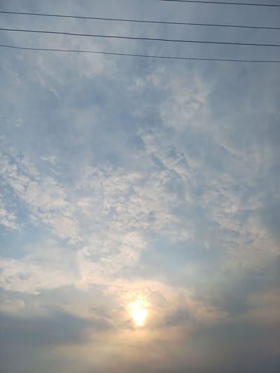 The image shows a sky with clouds and the sun. There are also power lines visible in the upper part of the image. The sun is partially obscured by clouds, creating a hazy glow. The clouds are varied in texture, with some appearing wispy and others more dense. The overall lighting suggests it might be late afternoon or early morning. There are no people or any other identifiable objects or structures within the frame. The location in Trypillia, Ukraine is not evident from the visual information provided in the image.