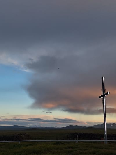 The image shows a landscape under a dramatic sky. Dark storm clouds dominate the left, contrasting with lighter, colorful clouds on the right near the horizon, possibly indicating sunset or sunrise. A metal structure resembling a cross is positioned near the top center of the frame. The landscape features green fields or vegetation, possibly a hillside, and what appears to be a mountain range in the distance. A fence line runs along the edge of the field. There is a bird in the sky. The location in the city of Anipemza, Armenia is not evident in the photo.