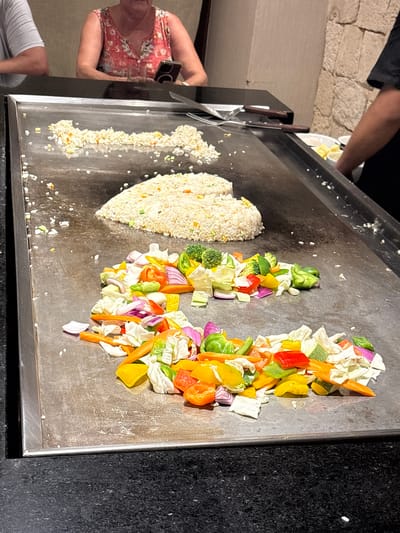 In the city of Punta Sam, Mexico, a hibachi grill is prepared for cooking. Two mounds of fried rice, one shaped like a heart, sit on the left side of the grill. In front of the rice, a colorful medley of chopped vegetables, including bell peppers, onions, carrots, and broccoli, is arranged in two piles. A person's arm, wearing a black shirt, is visible on the right, suggesting they are the chef. A woman with a floral top and a man wearing a grey shirt are seated at the table in the background, partially visible. The scene is set in a restaurant with a stone wall in the background.