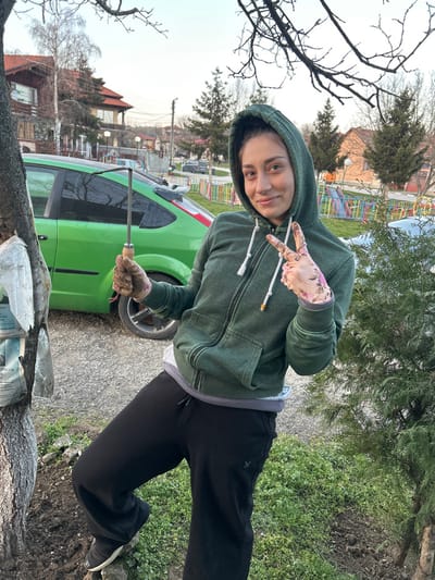 A female individual is depicted outdoors, posing for the camera in the city of Bukovlak, Bulgaria. She stands with her left foot slightly elevated, facing forward with a slight smile, looking directly at the camera. She wears a dark green hooded zipper jacket over a light undershirt, black sweatpants, and dark shoes. Her hands are gloved; the left hand, covered by a dark, soil-stained glove, holds a small metallic gardening hoe with a wooden handle. The right hand, covered by a floral-patterned, soil-stained glove, displays a two-finger "V" gesture. To the left, a tree trunk is visible, wrapped with several white, possibly paper or plastic, items, and surrounded by disturbed dark soil. In the mid-ground, a lime green Ford Focus hatchback is parked on a gravel surface. Behind the vehicle and to the right, a colorful children's playground structure with slides and climbing elements is partially visible behind a multi-colored fence. Residential buildings with varying roof styles and numerous trees, including bare deciduous and evergreen conifers, are present in the background under a pale sky.