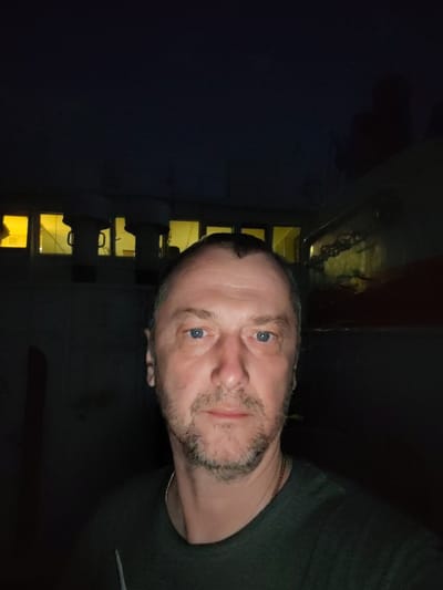 A dimly lit selfie of a middle-aged man with short, dark hair and a stubble beard is taken outdoors. The man has blue eyes and is wearing a dark green shirt and a gold chain. In the background are structures with illuminated windows, possibly a building. The location may be Ermolino, Russia.