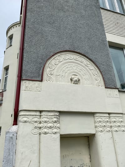 The image shows a close-up, exterior, eye-level view of a light-colored building facade with decorative elements in Sortavala, Russia. The building has a rough grey textured upper section and a smoother, lighter colored section below. A prominent feature is a semi-circular decorative relief above a doorway. This relief contains a central grotesque face, possibly a lion or dog, surrounded by concentric circular patterns and geometric designs. To the left of the grey section is a curved tower-like structure with small, square-topped windows. A dark red pipe runs vertically along the side of the building. The lower portion of the facade features a doorway and decorative pilasters with repeating oval and circular motifs. The building appears aged, with peeling paint visible on some surfaces. The overall architectural style suggests an Art Nouveau or similar early 20th-century influence.