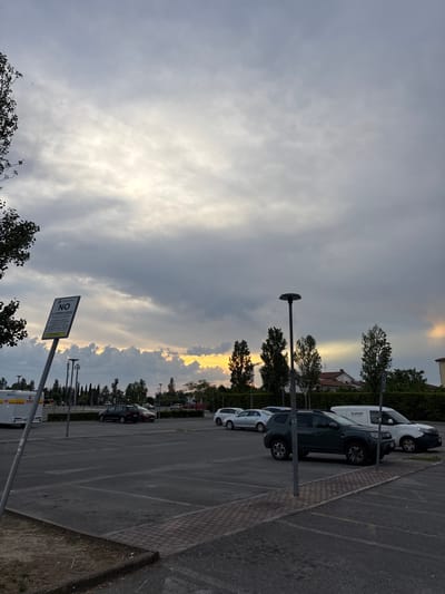 The image depicts a parking lot in Pisa, Italy. Several cars are parked along the right side of the frame. The sky is filled with clouds, with a patch of golden light breaking through near the horizon. In the foreground, there is a parking restriction sign. Streetlights are visible along the edge of the parking lot. Trees line the horizon behind the parked cars.