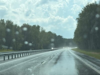 The image depicts a wet road with vehicles in the distance, viewed from inside a vehicle, possibly a car. The road appears to be a highway or a major road, with a guardrail visible on the left side. The weather is overcast, with clouds filling the sky, and there are water droplets on the camera lens, suggesting rain or recent rain. Trees line the sides of the road. The lighting conditions are dim. The scene is a generic road environment and does not contain any specific landmarks or features that would identify it as being located in the city of Tyumen, Russia.