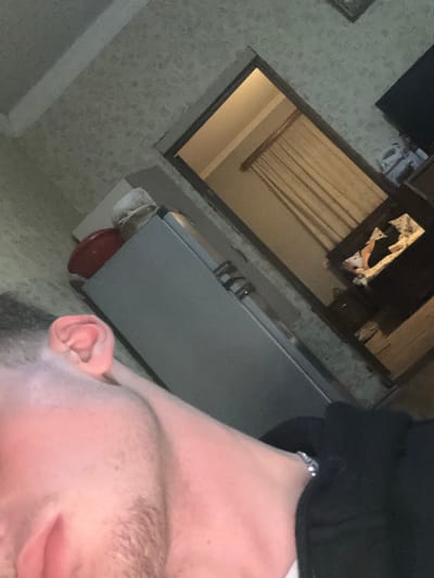 The image depicts an indoor scene in Durgena, Georgia, taken from a low-angle perspective. In the immediate foreground, the left side of a person's head, neck, and shoulder are visible. The person has fair skin, light-colored facial stubble along the jawline and chin, and is wearing a dark-colored garment that covers the shoulder and arm. A silver-colored link bracelet is present on the visible wrist.

The background shows a room with light-colored, patterned wallpaper. To the left, a light-toned rectangular object, possibly a small cabinet or table, holds a red bowl and a white, crinkled object on its surface. Adjacent to this, a large, light-grey, rectangular appliance, resembling a refrigerator or tall cabinet, is positioned with horizontal handles visible. Further into the background, an open doorway reveals a portion of an adjacent room. This room has light-colored walls and a window covered by light-toned vertical blinds or curtains. Below the window, a dark-colored piece of furniture, possibly a dresser or console, is present, with a white, patterned fabric and other indistinguishable items placed on its surface. The overall lighting appears to be interior artificial illumination.