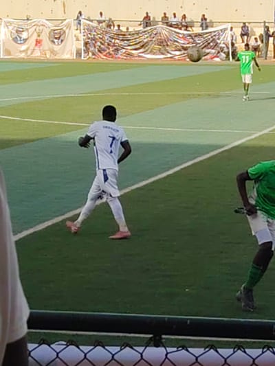 The image captures a daytime football match taking place on an artificial turf pitch, identified as being in Sudan. In the foreground, a player wearing a white jersey with the number "7" and the partially visible name "SHARION" on the back walks across the field, wearing distinctive pink football boots. To the right, another player in a green jersey with white shorts and dark boots is partially visible. Further down the field, closer to the goal, a player in a bright green jersey with the number "11" on the back stands. A football is prominently featured in mid-air near the goal, suggesting a moment of active play.

Behind the goal, a line of spectators is seated on a low, light-colored wall. Several banners or flags are draped across the back of the goal area; one banner clearly displays three circular portraits of individuals, though any accompanying text is illegible due to distance and resolution. The background suggests an outdoor stadium environment with clear, sunny weather. A dark mesh fence is visible in the bottom foreground, indicating the perspective of the photographer from behind a barrier, likely spectator seating.