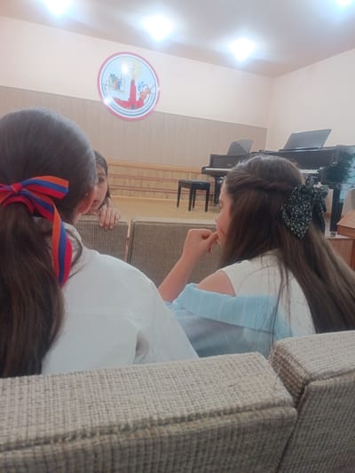 In Nor Hachin, Armenia, an indoor scene depicts four young girls sitting on cushioned benches. Two girls are in the foreground, facing away from the camera and towards a stage. One girl on the left has her back to the viewer, with dark hair tied in a ponytail adorned with a red, blue, and orange ribbon. The other girl on the right, also facing away from the camera, has long brown hair with a floral-patterned dark green bow. A third girl, partially visible between the two in the foreground, is looking towards the girl on the left. A fourth girl is seated on the right side of the image, facing left and looking down. The background features a stage with a black grand piano and a small stool, and a circular emblem is mounted on the wall. The lighting appears to be from overhead fluorescent lights.