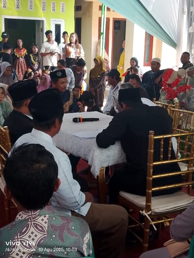 The image depicts a daytime social gathering or ceremony occurring in Jagapura Kulon, Indonesia. The central focus is a group of six men seated around a rectangular table draped with a white, ruffled tablecloth, using gold-colored Chivari-style chairs. A black microphone with an orange band and white papers are prominently placed on the table. Two men in the immediate foreground are seen from behind, one wearing a patterned green batik shirt and the other a light blue collared shirt with brown pants. On the left side of the table, a man adorned in a black peci and an elaborate dark blue traditional garment with a distinctive headdress appears to be engaged in conversation or leading a discussion. Another man in a black peci is seated beside him. Opposite them, a man in a black shirt is partially visible from behind, and a man in a white peci and blue patterned long-sleeved shirt is seated, holding what appears to be a stick or cane.
In the mid-ground and background, an estimated twenty to thirty additional individuals of various ages are present, standing and observing the central activity. This larger group includes individuals dressed in traditional Indonesian attire, such as women wearing hijabs and men in peci. Distinct figures include a young woman in a red and gold traditional dress with an ornate collar and headband, and another young girl in a green and gold traditional outfit, both positioned on the far left. A woman holding a baby is visible in the right background. The environment features a light green fabric canopy extending overhead to the upper right. Architectural elements in the background include a light green building with a white-trimmed doorway on the left and another building with reddish-brown elements on the right. Red artificial flowers and garlands are arranged on a decorative stand behind the seated men on the right. The ground in the foreground is covered with a red carpet or matting.