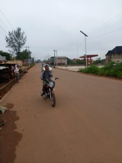 The image depicts a man riding a motorcycle on a paved road in Ita Oniyan, Nigeria (coordinates: latitude: 7.295, longitude: 5.159). The road is brown and dusty, with a small ditch to the left. The man is wearing a blue shirt and black pants and appears to be talking on a phone. He is riding towards the camera. There are several buildings in the background, including a gas station on the right and a few residential buildings. Several power lines run across the sky. The weather appears overcast with a muted grey sky. The trees on the left side of the road are sparse and green.