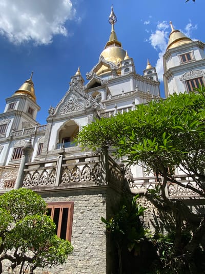 The image captures a low-angle shot of the Buu Long Pagoda located in Dĩ An, Vietnam. The pagoda features a white exterior with ornate carvings and golden spire tops. The bright blue sky with scattered white clouds is visible between the pagoda structures. Parts of the pagoda are obscured by lush green trees. The exterior of the building is constructed from brick with various window openings.