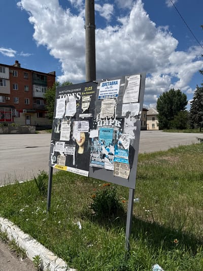 The image shows an outdoor bulletin board covered in various advertisements and notices, standing in a grassy area with a concrete curb in the foreground. Behind the bulletin board, a paved area, likely a parking lot or plaza, is visible, with buildings and trees in the background. The sky is blue with scattered white clouds.
The bulletin board displays numerous pieces of paper, many of which are torn or weathered. Some visible text on the board includes "ДОСТАВКА", "РАБОТА", "РЕМОНТ ХОЛОДИЛЬНИКОВ", "МАТЕРИНСКИЙ КАПИТАЛ", and "ДЕНЬГИ ПЕНСИОНЕРАМ".
The environment suggests a public outdoor space in a town or city. The buildings in the background appear to be residential or commercial structures. The presence of trees and grass indicates a potentially suburban or semi-urban setting.
The time of day appears to be daytime, given the lighting and the blue sky. No people are directly visible in the frame, but the bulletin board implies a public area where people would pass by. The overall scene is a typical snapshot of a community bulletin board in a Russian locale.