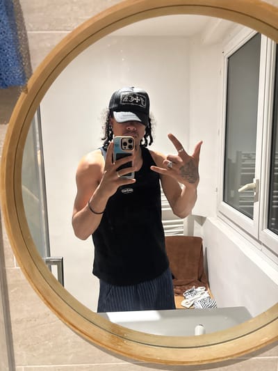 A person is taking a mirror selfie in what appears to be a bathroom. The person is wearing a black tank top, striped pants, and a black and white baseball cap. They are holding an iPhone in their right hand and making a hand gesture with their left hand. The bathroom features a round mirror with a gold frame, white walls, beige tiles, a window, and what appears to be a towel rack. There is no explicit evidence within the photograph to confirm the location in Massy, France.