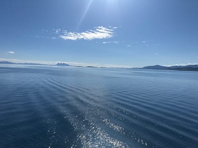 This is a photograph of a body of water, likely a fjord, with mountains in the distance. The sky is blue with a few scattered clouds. The sun is shining brightly, creating reflections on the water. The image appears to have been taken from a boat or other vessel, as there are visible ripples on the water's surface, suggesting movement. The distant landmasses are hazy and appear to be mountainous. The scene suggests a clear, sunny day in a coastal or maritime environment. The location is specified as Lødingen, Norway.