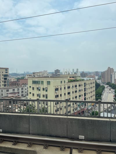 An elevated shot captures a cityscape in Dongguan, China, under a cloudy sky. In the foreground, railway tracks run horizontally across the bottom of the frame. Above them, a concrete barrier is visible, followed by a decorative metal railing. Beyond the railing, a street lined with trees and parked cars stretches into the distance. Buildings of varying heights and architectural styles populate the scene, some with visible air conditioning units on their exteriors. The overall impression is one of urban development with elements of everyday city life.