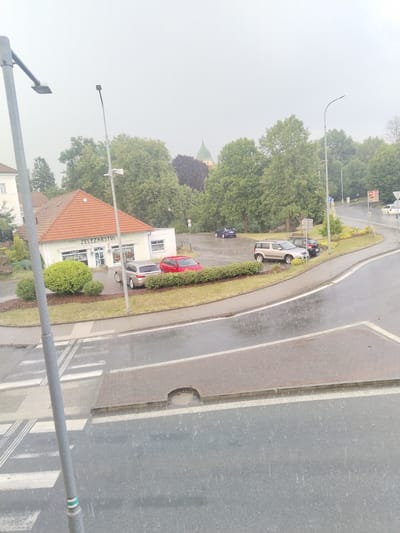 The image depicts a street scene in Častolovice, Czechia, during a rain shower. The foreground features a wet asphalt road with white lane markings and a pedestrian crossing. A grey lamppost stands to the left, partially obscuring a section of the crossing. In the mid-ground, a small commercial building with a red tiled roof is visible, displaying the text "ZELEZARSTVI" above its entrance, indicating it is a hardware store. A silver car and a red car are parked in front of the store. Beyond the building, a row of lush green trees lines the landscape. Further in the background, more trees and the indistinct spire of a building can be seen under a cloudy, overcast sky. The rain is evident from the streaks on the road and the droplets in the air. The overall atmosphere is gloomy due to the weather conditions. There are no people visible in the scene.