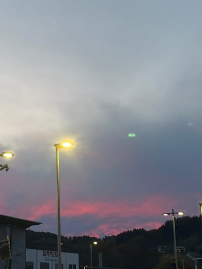 This image depicts an evening or dusk scene in Oppenau, Germany. The sky is largely overcast, featuring a prominent horizontal band of vibrant pink and red clouds stretching across the lower portion, transitioning to purple and grey tones higher up. A faint, diffuse green light anomaly, shaped somewhat like a butterfly or two interconnected ellipses, is visible in the upper-middle right quadrant of the sky.

In the foreground, from left to right:
- The upper section of a building with a light-colored, possibly corrugated metal, facade is visible on the far left. A sign on this building displays the text "BIMMERLE" and "EDELBRÄNDE," identifying the location as the Bimmerle KG distillery in Oppenau.
- A partially obscured streetlight with a bright yellow-orange glow is present on the far left, behind some dark tree foliage.
- A tall, slender streetlight stands in the left-center, emitting a strong yellow-orange light with a visible lens flare.
- Further to the right, two additional streetlights with similar yellow-orange illumination are visible, set against the backdrop of the sky and hills.

The background consists of dark, densely forested hills that rise against the horizon on the right side of the image, silhouetted beneath the colorful sky. No individuals or specific actions are discernible within the frame.