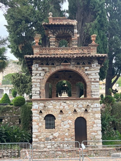 This image captures a picturesque view of the small, ornate villa located within the gardens of Villa Comunale in Taormina, Italy. The villa, constructed from a mix of stone and brick, features multiple levels and arched openings. The lower level has a dark wooden door and a arched window with a metal grate. Above this, there are two levels with open arches supported by brick pillars. The structure is topped with a tiled roof and decorative elements. The villa is surrounded by lush greenery, including tall trees, manicured hedges, and flowering plants. In the foreground, a metal fence encloses the area. The sky appears overcast, contributing to the soft lighting of the scene.