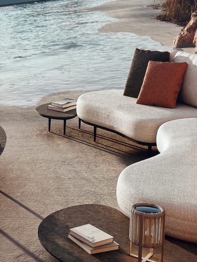 The image depicts an outdoor seating area on a sandy surface, possibly a beach or a meticulously designed patio that mimics a beach environment. The scene is set adjacent to calm, shimmering water. In the foreground, there are two round, dark-colored coffee tables. The table on the left has two books stacked on it. The table on the right holds a cylindrical, wicker-like lamp with a blue illuminated top, supported by thin legs. Behind the tables, a large, modular outdoor sofa is visible. The sofa is upholstered in a light-colored, textured fabric and features a curved, organic design. It is adorned with two large throw pillows: one dark brown and textured, and another in a vibrant rust orange, also textured. The backdrop shows the gentle lapping of water against a sandy shore, with sparse dry vegetation and rocks to the right. The lighting suggests a daytime setting, with soft shadows cast on the ground. The image does not contain any elements or context that would suggest a location in the city of Mol, Belgium.
