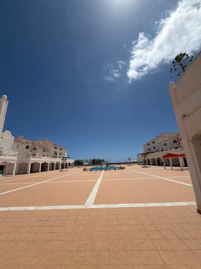 The image depicts a view of a public square in Arroyo de la Miel-Benalmádena Costa, Spain. The square is paved with reddish-brown tiles marked with white lines. Flanking the square are white buildings with balconies and arched supports. In the distance, there is a glimpse of a swimming pool with trees and a distant view of the sea. The sky is blue with some scattered white clouds. The sun is visible in the lower left.