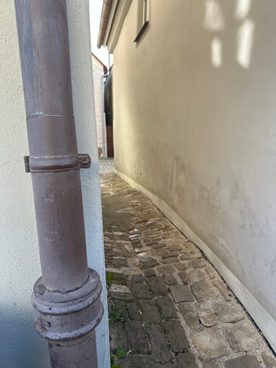 This image depicts a narrow, cobblestone alleyway in Weißenburg in Bayern, Germany. The scene is illuminated by natural daylight, casting distinct shadows. On the immediate left, a weathered, dark brown metal downspout is vertically affixed to a light-colored building wall. This downspout features a securing bracket approximately midway up the visible section and a decorative, fluted base near the bottom. The wall it is attached to is painted a light beige and shows minor discoloration.
To the right, another building wall, also light beige, extends into the background, curving slightly to the left. A small rectangular window with a dark frame is visible high on this wall. Below the window, multiple rectangular shadows, consistent with windowpanes or an adjacent architectural feature, are projected onto the wall surface. The lower section of this wall exhibits signs of dampness or age-related discoloration. A dark-colored roof gutter system is visible along the roofline above this wall.
The ground surface of the alley consists of an uneven paving of irregular, multi-tonal grey and beige cobblestones. Small patches of green vegetation are observed growing between the cobblestones, particularly on the left side of the path. The alley narrows considerably into the background, with a dark, obscured passage visible at the farthest point. No individuals, animals, or active events are present in the frame.