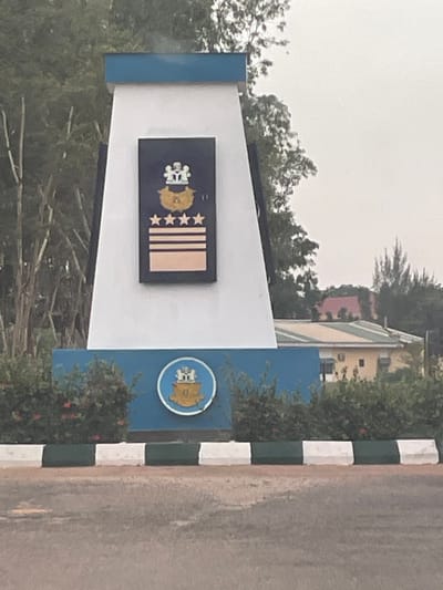 The image shows a sign at the Nigerian Defence Academy in Afaka, Nigeria. The sign features the Academy's emblem and rank insignia. The environment includes trees, shrubs, a building in the background, and a striped curb. The image appears to be taken during the day under overcast conditions. There are no people visible in the frame.