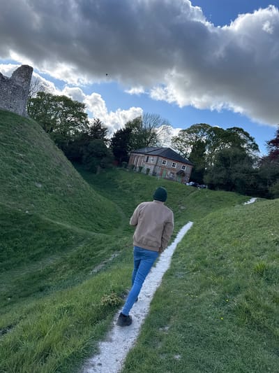 The image captures a scenic outdoor view, predominantly featuring a person walking up a winding, light-colored path across a grassy hillside. The path, possibly made of chalk or gravel, cuts through vibrant green grass that is speckled with small yellow flowers in places.
The main subject, seen from behind, is wearing a light brown or beige jacket, blue jeans, and a dark green beanie. They are positioned on the path, heading towards the middle-left of the frame.
In the middle ground, set among dense green trees, stands a rustic stone and brick house with a brown roof and white-framed windows. Two dark-colored cars are faintly visible parked near the house, partially obscured by the foliage. To the far left and lower part of the frame, there are weathered stone ruins, suggesting an ancient structure or a historical site.
The sky above is a dynamic mix of bright blue patches and large, dramatic grey and white clouds, indicating a partially cloudy day. A small, dark bird is captured mid-flight against the cloudy expanse in the upper left section of the image.
The overall scene suggests a peaceful stroll or exploration of a natural and historical landscape, likely in the countryside of King's Lynn and West Norfolk, United Kingdom. The time of day appears to be daytime, with ample natural light illuminating the scene. There is no visible text in the image.