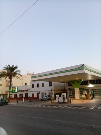 The image shows a street scene in Olula del Río, Spain, with a gas station and a restaurant prominently featured. A palm tree stands to the left of a building with a sign that reads "RESTAURANTE". The gas station has a white canopy with green trim, supported by green pillars. Several cars are visible, including a silver hatchback parked near the restaurant and a dark green car parked on the street. The sky is clear and pale blue, suggesting either early morning or late evening. The scene appears quiet, with no people clearly visible engaging in activity. The road in the foreground has pedestrian crossings.
