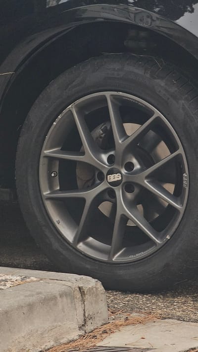 The image shows a close-up of a car wheel and tire parked next to a concrete curb in Zaragoza, Spain. The car is dark-colored, possibly black, and the wheel is a multi-spoke alloy design in a dark grey finish. A prominent "BBS" logo is visible in the center of the wheel. The tire appears to be worn. The concrete curb in the foreground is damaged and cracked, with pine needles scattered on and around it. The ground surface appears to be asphalt or a similar rough texture. The overall scene suggests a parking area, likely on a street or lot. There are no people or animals visible in the image. The lighting is natural, indicating daylight.