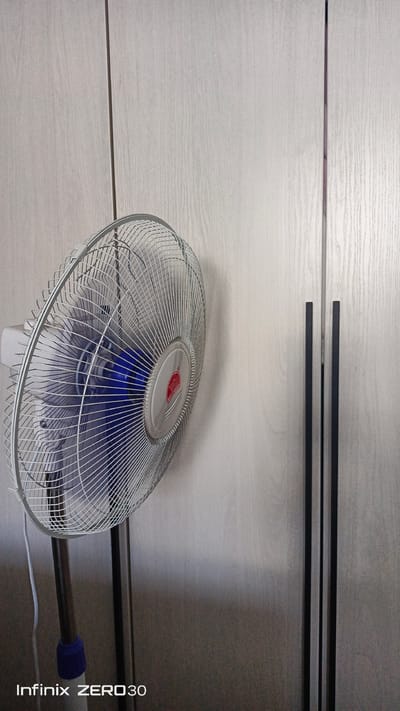 A standing fan with a silver grill and blue blades is positioned to the left of the frame, angled towards the right. The fan is on a dark pole with a blue attachment near the base. Behind the fan is a light-colored, wood-grain textured wall, likely a closet door or paneling, with two vertical black handles in the center right of the image. The bottom left corner of the image contains a watermark that reads "Infinix ZERO 30". The scene appears to be indoors, in a domestic setting. There are no people or animals visible in the image. The lighting is soft and diffused, suggesting an indoor light source or an overcast day. The image appears to be taken from a slightly low angle, looking up at the fan. There is no information available that can associate this image with the city of Bugulma, Russia.