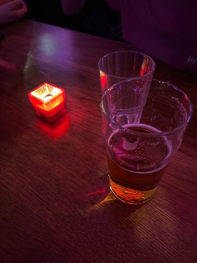 An overhead, slightly tilted shot captures two clear drinking glasses and a small red light source on a dark wooden table in a dimly lit indoor setting. The atmosphere is colored by strong red and purple/pink lighting, casting vibrant reflections and shadows.
In the upper part of the image, two transparent glasses lie horizontally. The glass on the right contains a small amount of amber liquid, likely beer, and has remnants of foam clinging to its inner walls, suggesting it was recently drunk from. This glass reflects the ambient red and purple light intensely. The glass on the left is empty and also reflects the colored lights.
Centrally placed on the table is a small, hexagonal or multi-faceted red object with a glowing internal light, possibly an LED tea light or decorative lamp, which casts a bright red glow onto the wooden surface around it.
The table itself is made of dark, reddish-brown wood with visible grain, reflecting the dramatic lighting. Blurry elements at the top left and bottom left edges hint at the presence of people, with a glimpse of clothing and what appears to be a hand or arm. No specific activity is shown beyond the presence of drinks, implying a relaxed social gathering in the evening or night. There is no visible text. The scene conveys a cozy, intimate ambiance, likely in a bar or restaurant in Ixelles, Belgium.