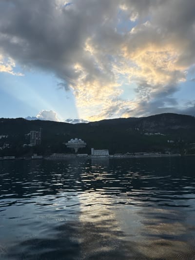 The image captures a shoreline at Гаспринский поселковый совет, Ukraine. A dark, mountainous landscape runs vertically along the right side of the frame, punctuated by buildings. The sky on the left is a blend of clouds and blue sky with sun rays beaming through. The body of water to the right reflects the light and sky.