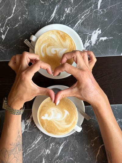 Two white cups of coffee with latte art are placed on a dark marble surface. Two hands are positioned between the cups, with fingers interlocked to form a heart shape. The hands are light-skinned, with manicured fingernails. One hand has a silver and gold watch on its wrist and a tattoo of a flower on its forearm. The scene appears to be set in a cafe or restaurant, suggested by the table setting and the presence of coffee cups and saucers. The lighting suggests daytime, possibly indoors due to the consistent illumination. The activity depicted is a gesture of affection or connection, using the coffee cups as a backdrop.